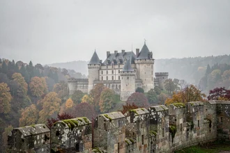 Château fort médiéval français dominant une vallée brumeuse entourée de forêts aux couleurs d'automne